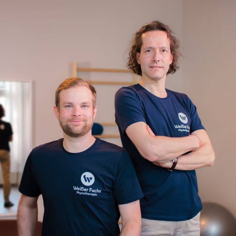 Ein Porträtfoto von den Physiotherapeuten Niklas Witt und Andreas Voß in einem Behandlunsgraum. Beide tragen schwarze T-Shirts. Andreas Voß stehend mit verschränkten Armen. Niklas Witt in der Hocke.